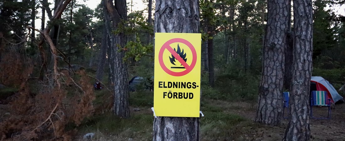 Tall med gul skylt med texten "ELDNINGSFÖRBUD" på. I bakgrunden syns ett tält.