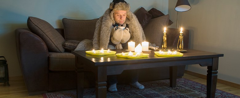 Manlig person sitter i en soffa inomhus och med ytterkläder på sig och fryser. På soffbordet finns tända stearinljus.