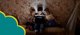 Tre barn sitter i en säng i ett mörkt rum och läser på en läsplatta. 