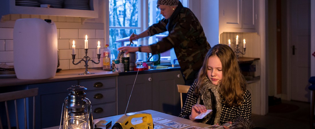 Vuxen och barn i ett kök utan ström. På köksbordet ligger en ficklampa. Tända ljus och vattendunk i bakgrunden.