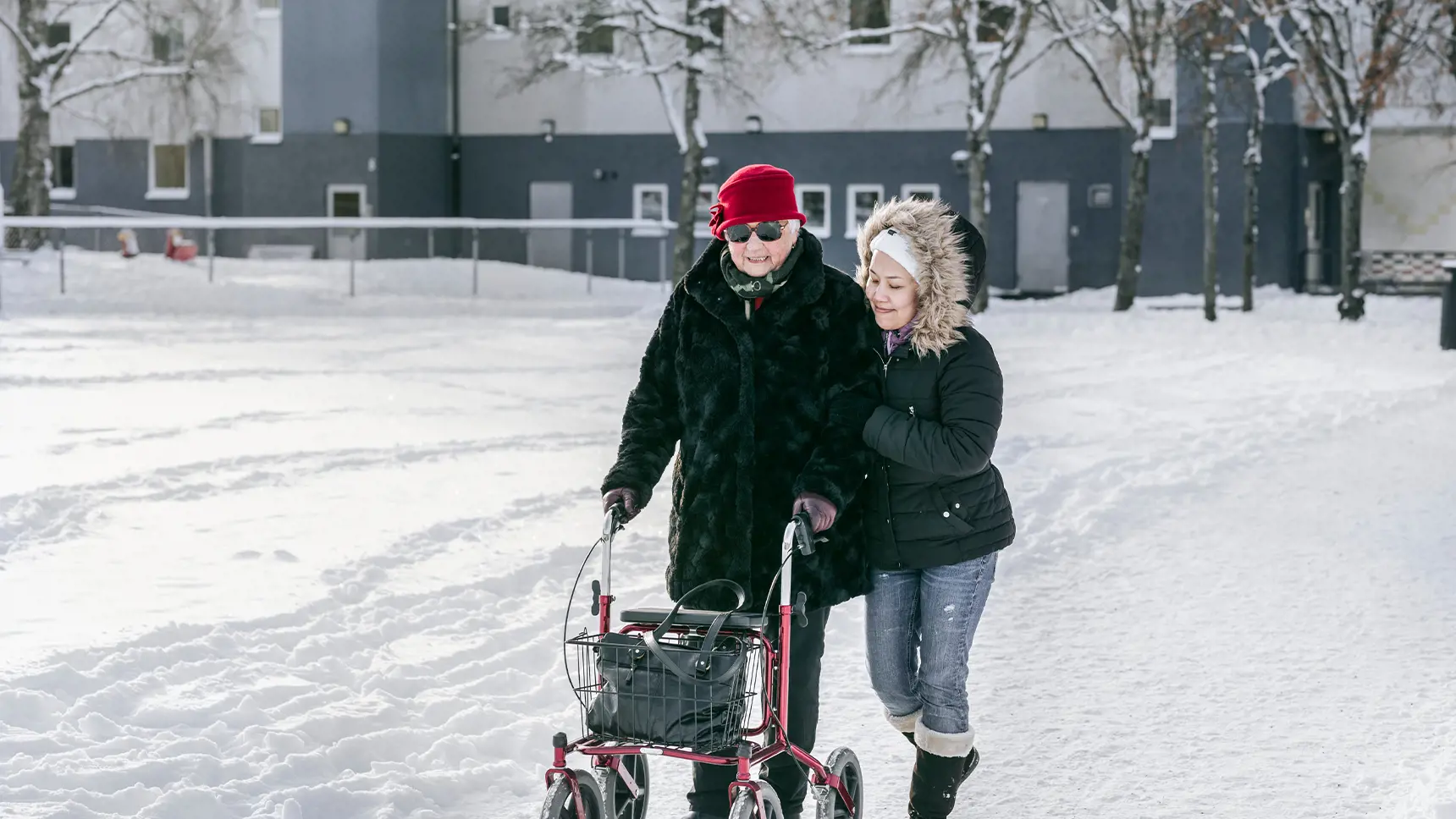 Ung flicka hjälper äldre dam med rullator genom snön.