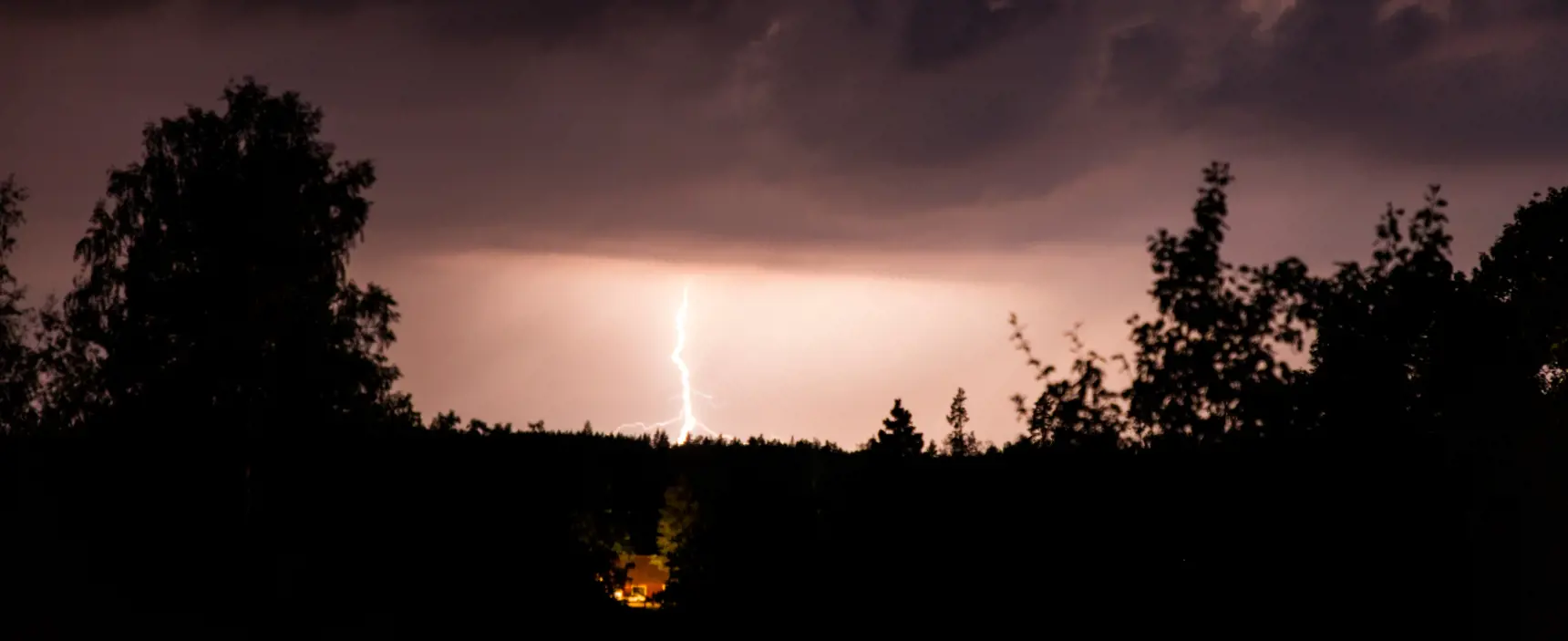Lightning striking a forest at night. The lightning illuminates a small red cottage.