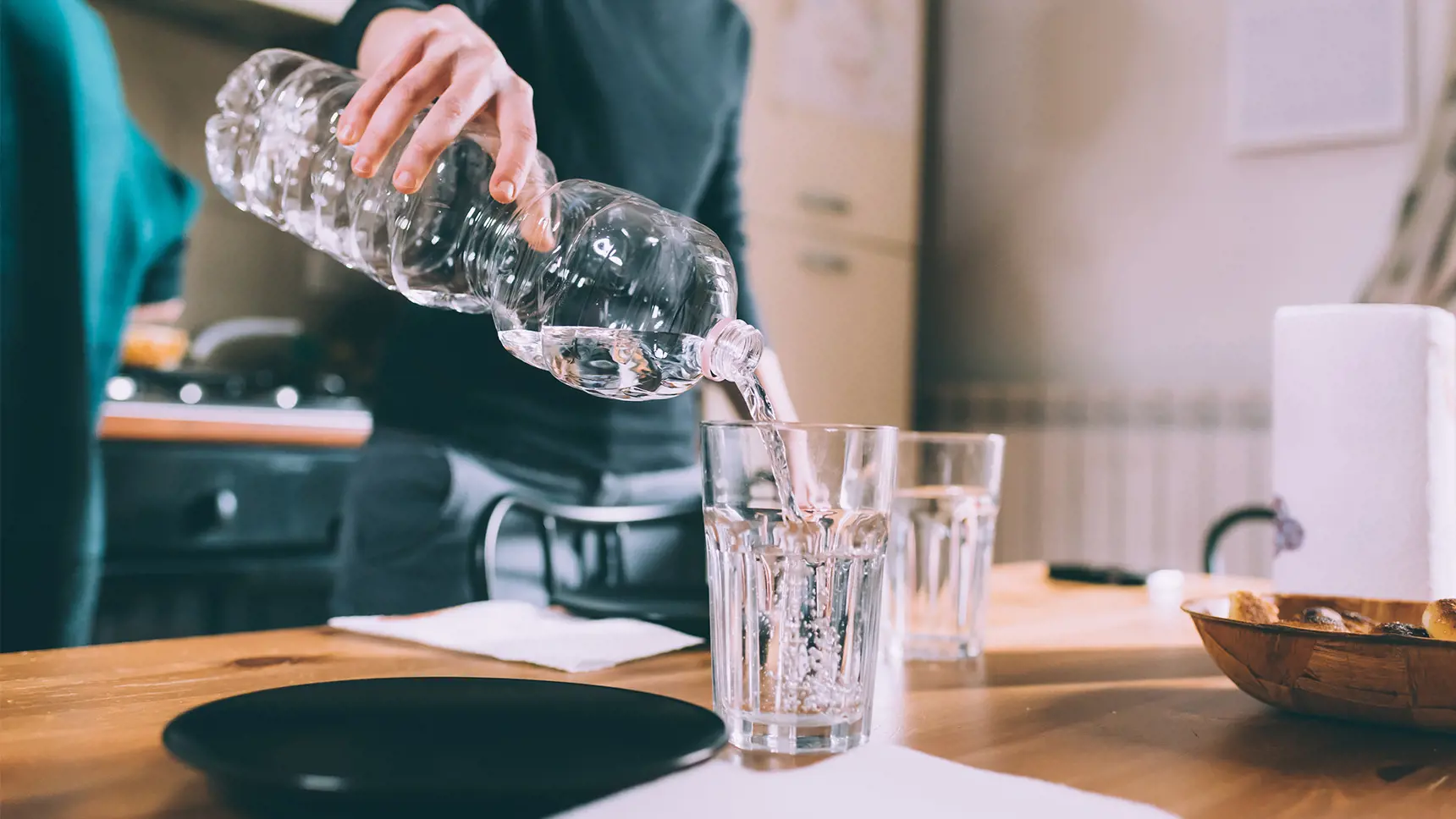 En person håller i en petflaska och häller vatten i ett glas som står på ett köksbord.