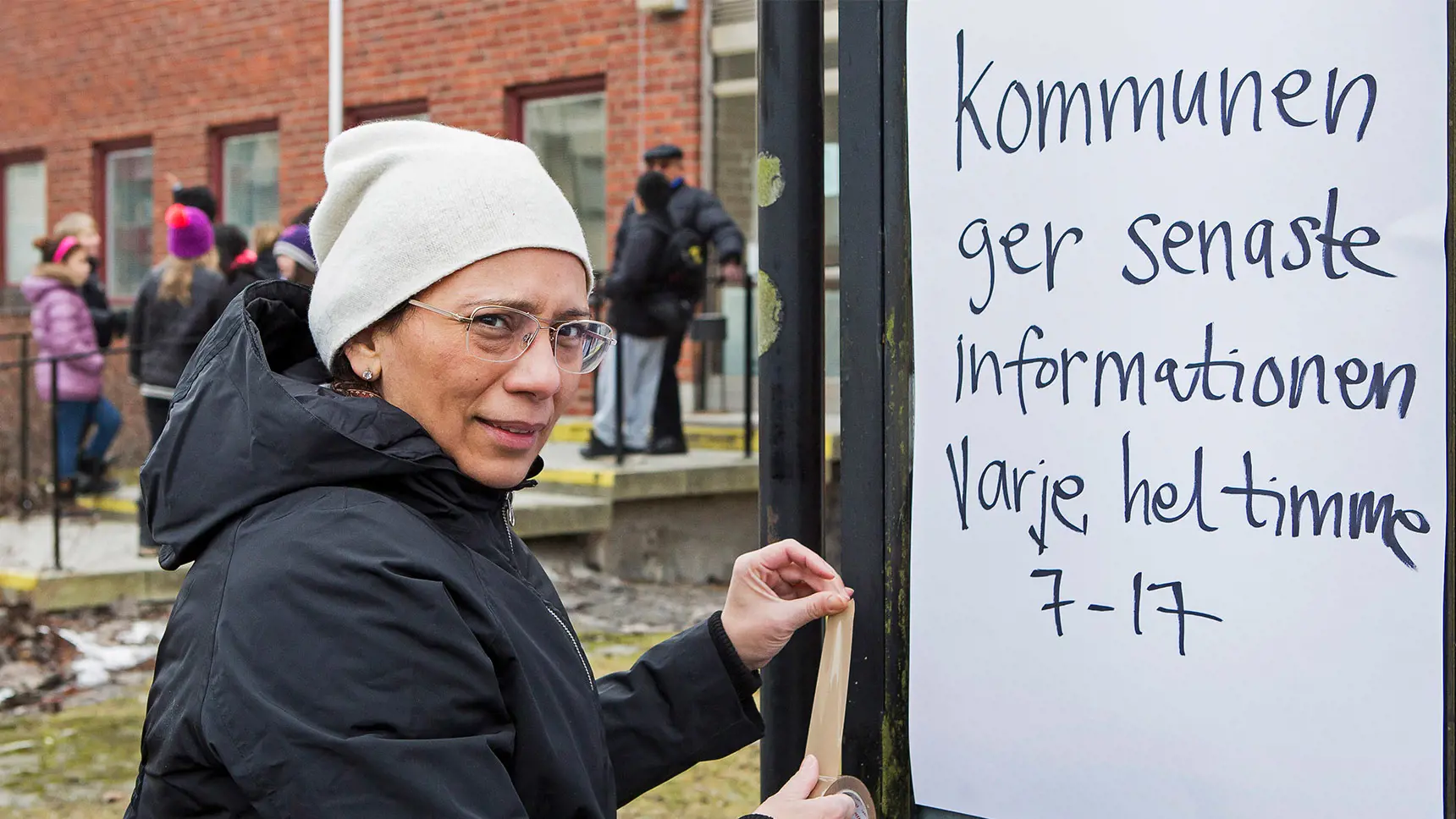 Kvinna står vid en anslagstavla med en tejprulle i handen. På anslagstavlan står det "Kommunen ger senaste informationen varje heltimme 7-17". I bakgrunden syns en grupp människor.