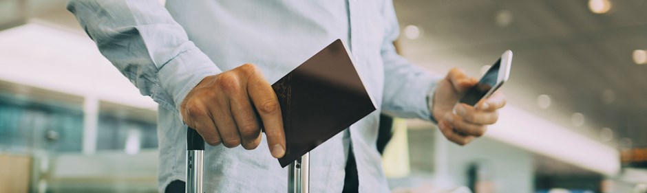 Resenär på flygplats med EU-pass och mobiltelefon i handen. Resväska skymtar. 