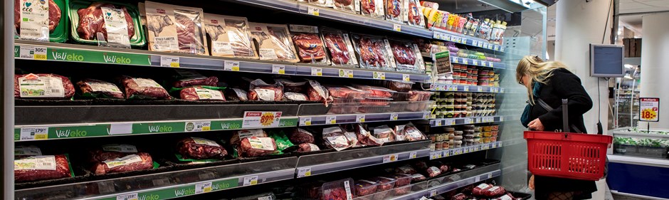 Hylla med köttprodukter i en livsmedelsbutik. Kvinna med korg som handlar.