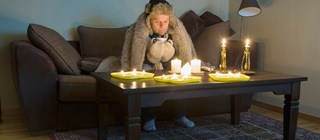 Manlig person sitter i en soffa inomhus och med ytterkläder på sig och fryser. På soffbordet finns tända stearinljus.