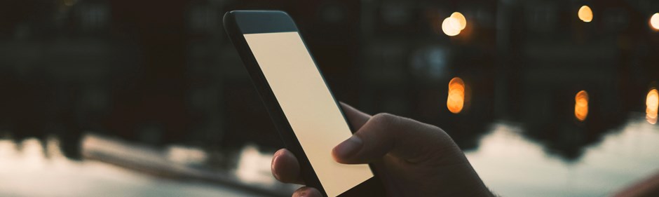Hand håller i mobiltelefon i kvällsljus med vatten och hus i bakgrunden.