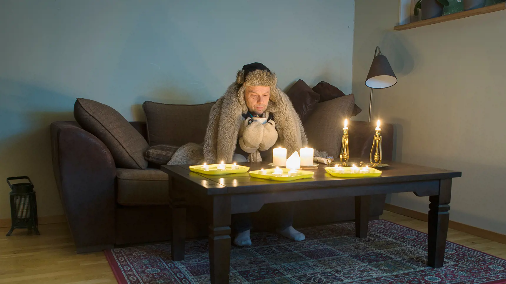 Man sitter i en soffa iklädd vinterkläder. På bordet framför honom är flera värmeljus tända.
