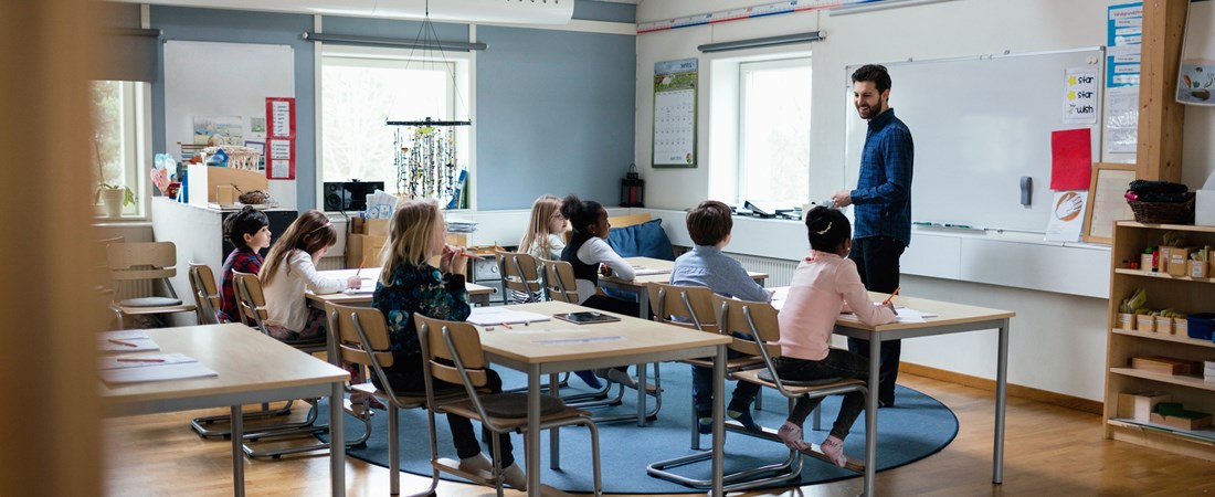 En lärare står framme vid tavlan och ler mot sina elever i klassrummet.