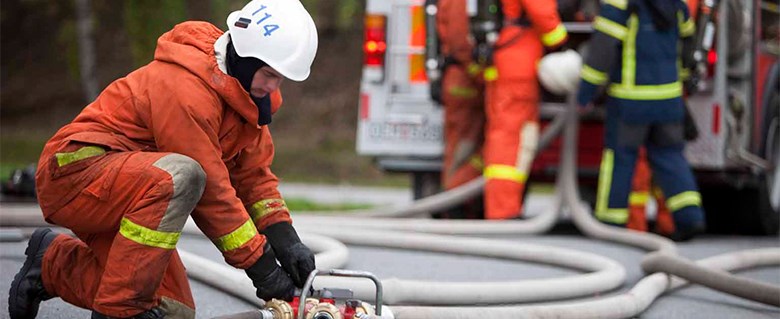 En orangeklädd brandman arbetar med en brandslang på marken.