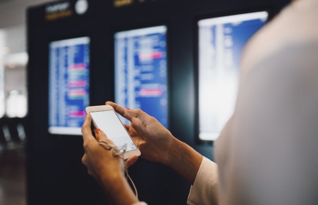 Person håller i en vit mobiltelefon framför informationsskyltar på en flygplats. 