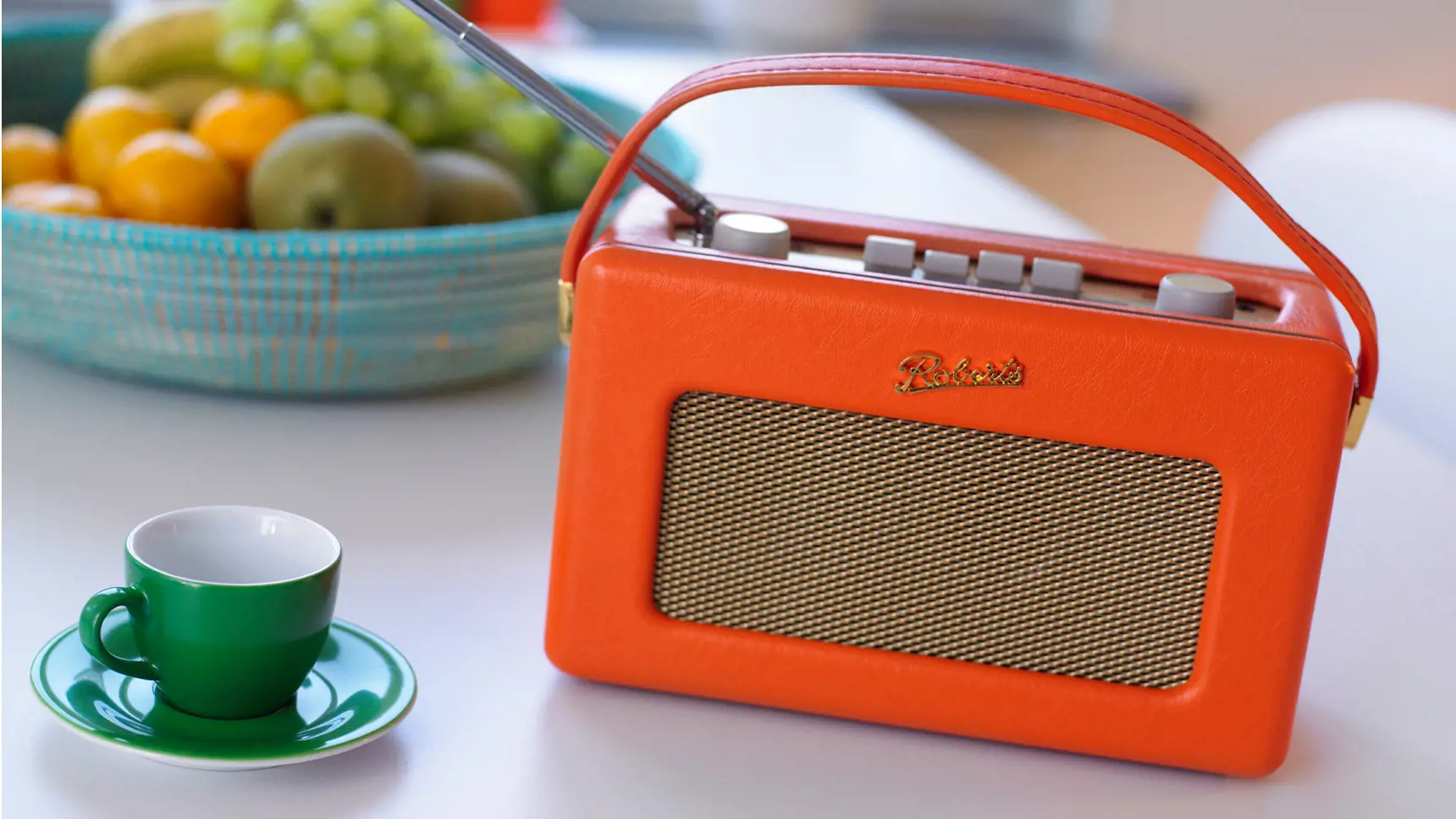 An old battery-powered radio on a table. Next to it is a coffee cup and a fruit bowl.